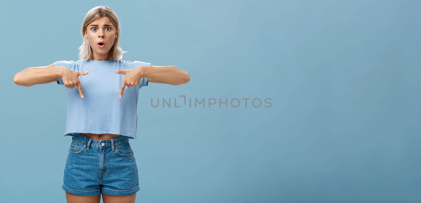 Lifestyle. Studio shot of worried caucasian young woman feeling nervous her shoes do not fit frowning asking advice nervously pointing down shocked while frowning and standing over blue background.