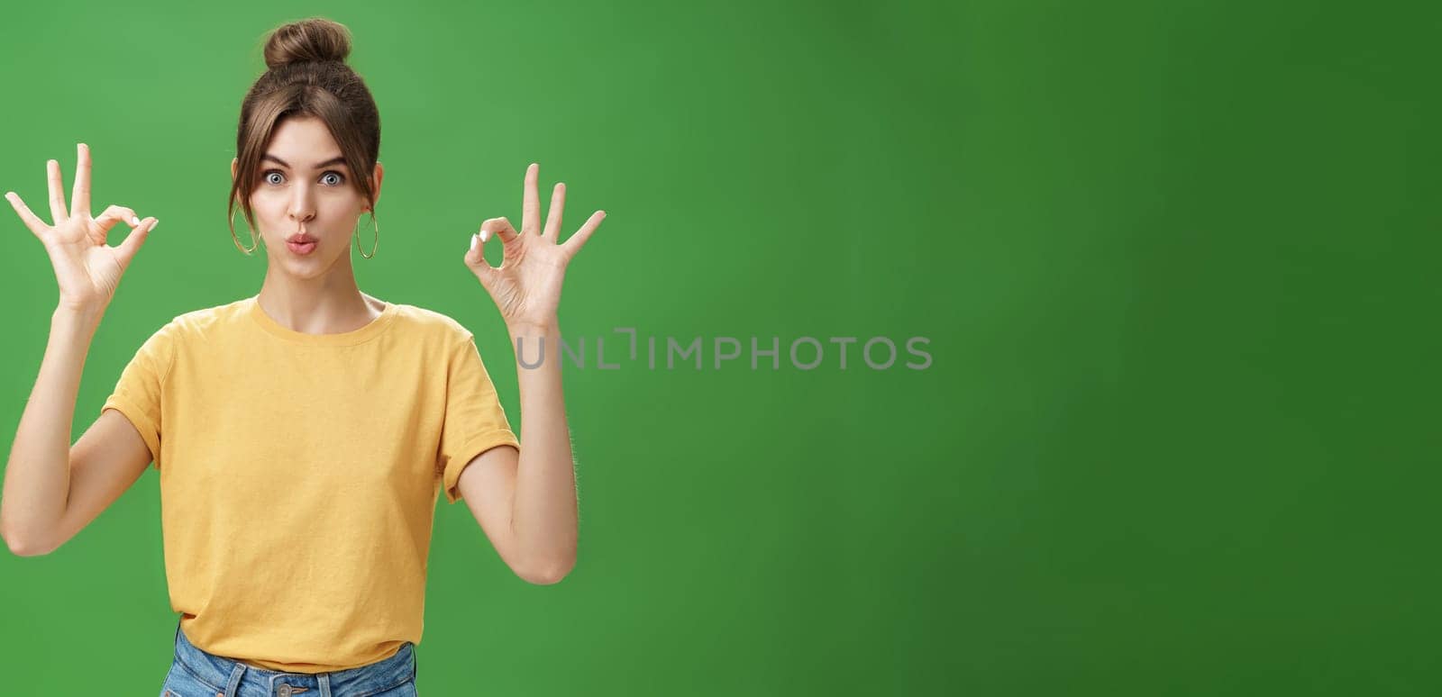 Portrait of silly cute and beautiful caucasian woman with combed hair in round earrings and yellow t-shirt showing okay, no problem gesture folding lips, reaffirming boss job goes well over green wall.