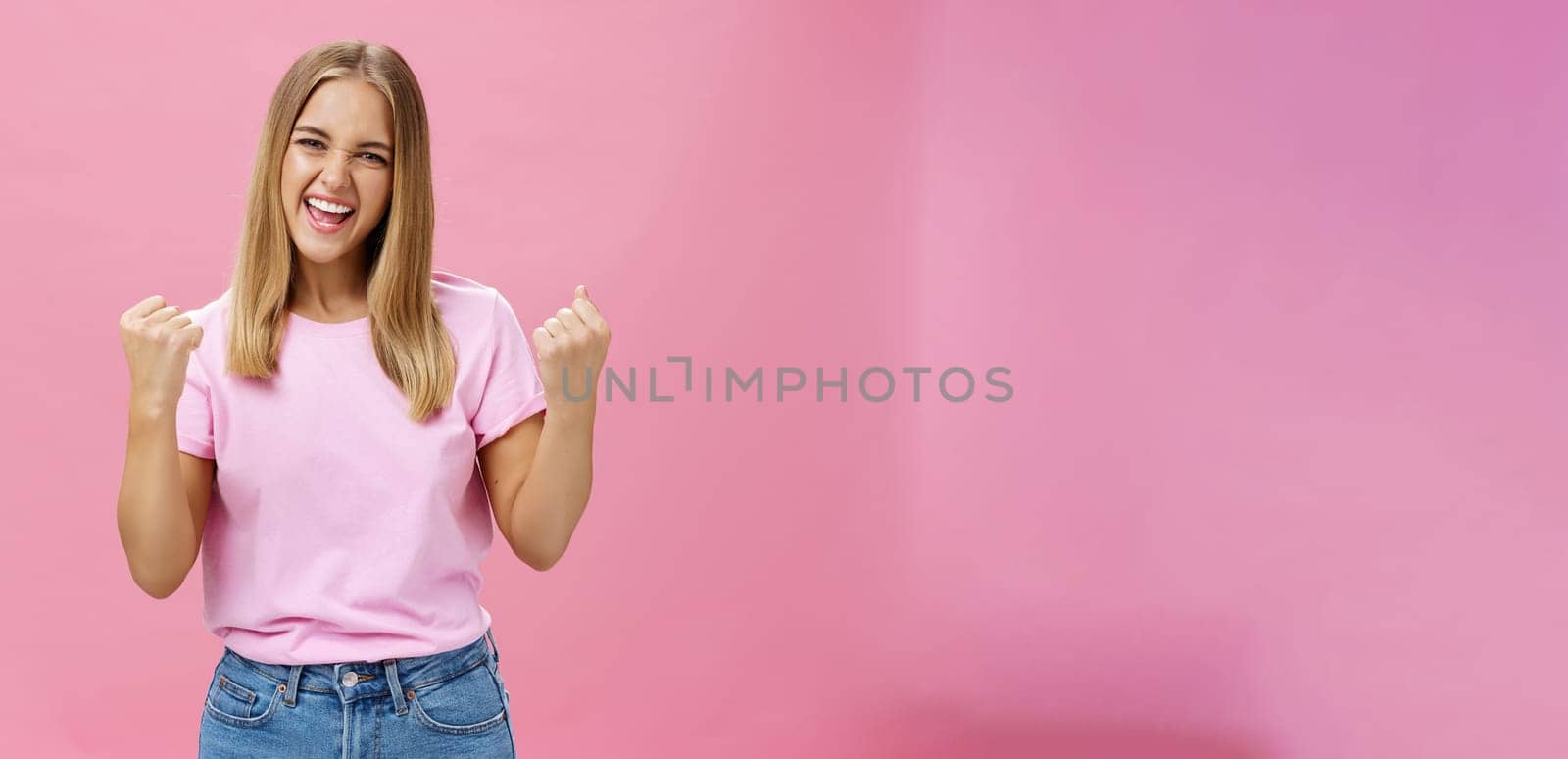 Yes I did it. Portrait of happy delighted successful young female student receiving award triumphing being excited and joyful raising clenched fists in cheer, gazing confident at camera over pink wall by Benzoix