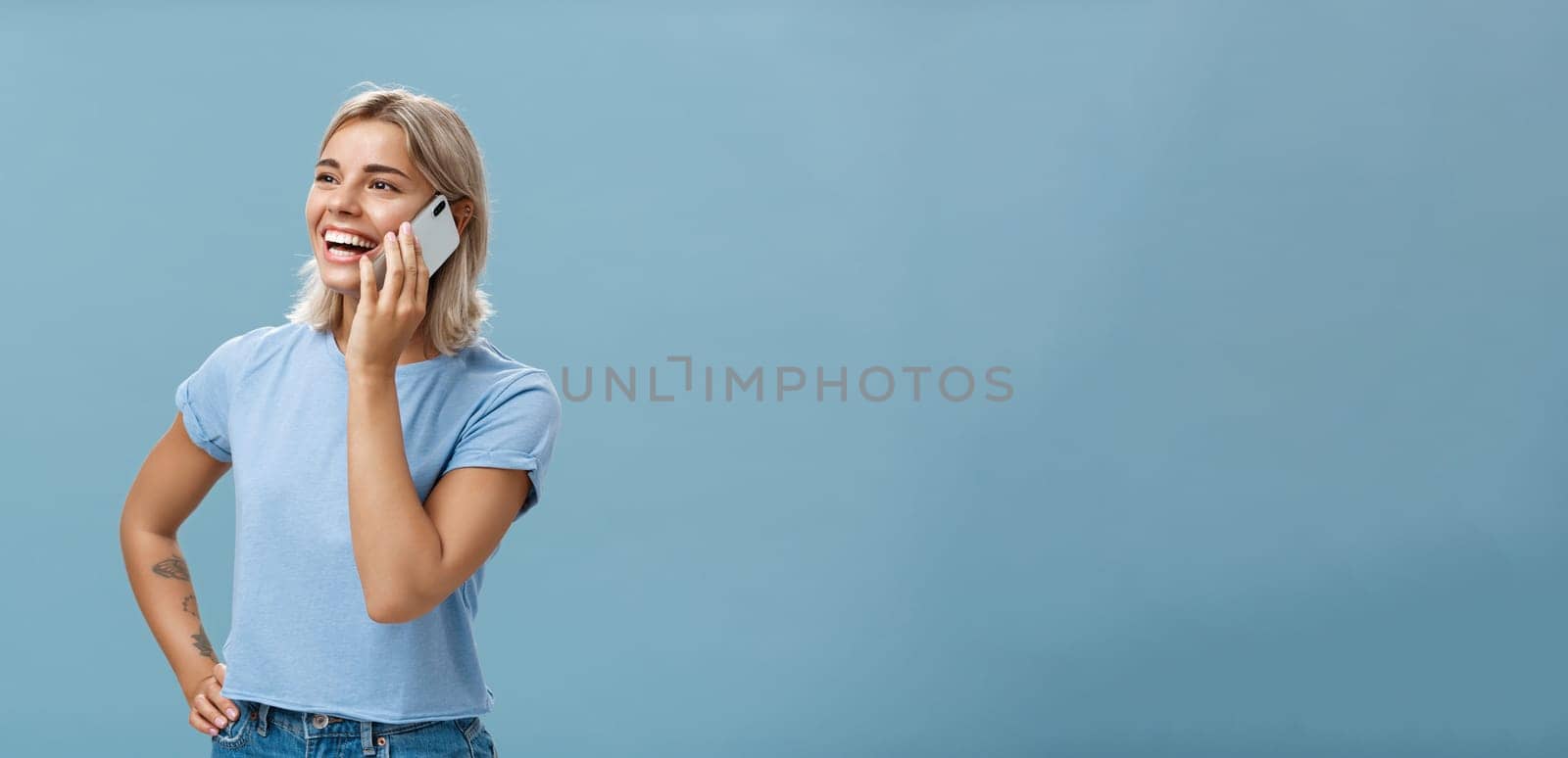 Lifestyle. Waist-up shot of sociable amused and happy attractive caucasian fair-haired woman in casual t-shirt standing half-turned gazing left with hand on hip while talking on smartphone over blue background.