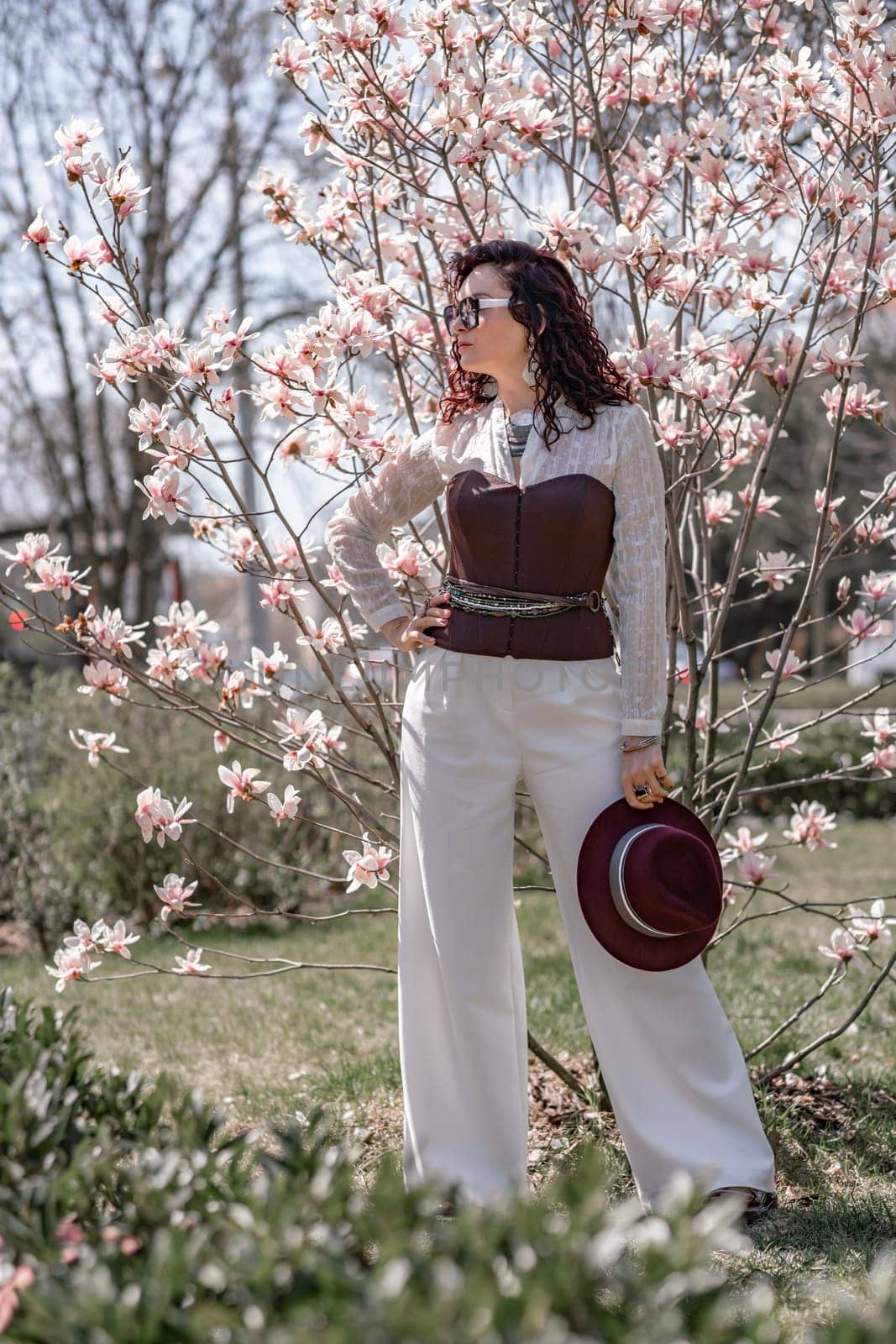 Magnolia park woman. Stylish woman in a hat stands near the magnolia bush in the park. Dressed in white corset pants and posing for the camera