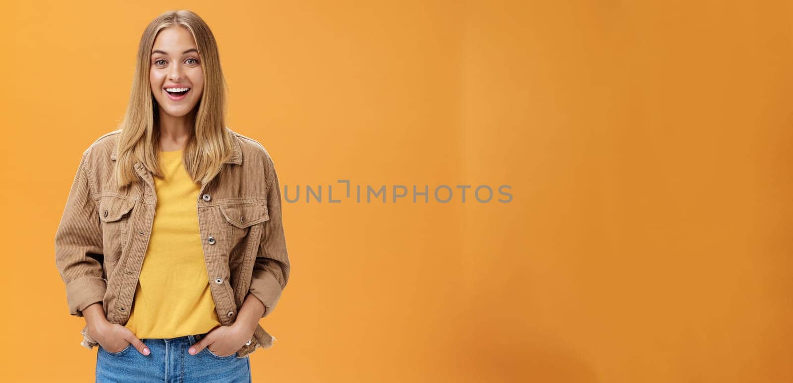 Charismatic tanned woman in corduroy jacket and yellow t-shirt ready for chilly autumn walk with friends smiling joyfully gazing entertained at camera holding hand in pockets casually over orange wall by Benzoix