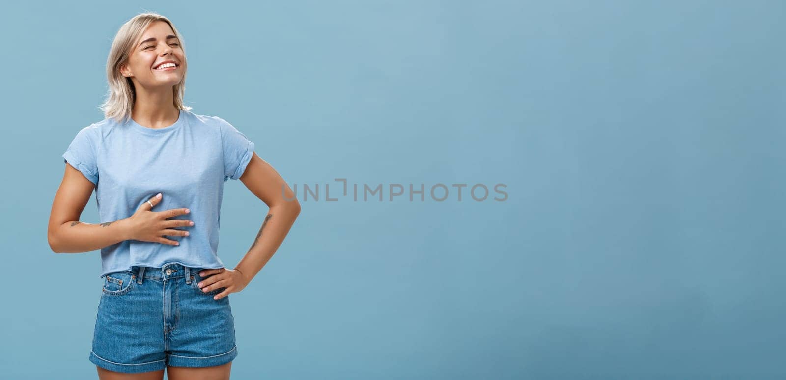 Cute girl likes eat after workout being pleased and stuffed after dinner. Portrait of satisfied attractive blonde young woman touching belly raising head up with broad smile over blue background by Benzoix