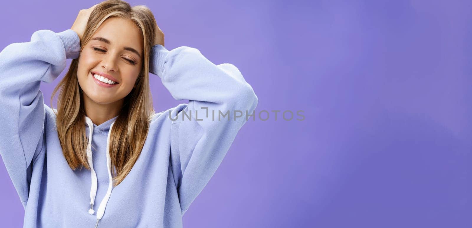 Tender relaxed charming woman with blond hair touching head joyfully smiling broadly with closed eyes daydreaming posing in trendy hoodie against purple wall finally getting rid of acne and pimples. Lifestyle.