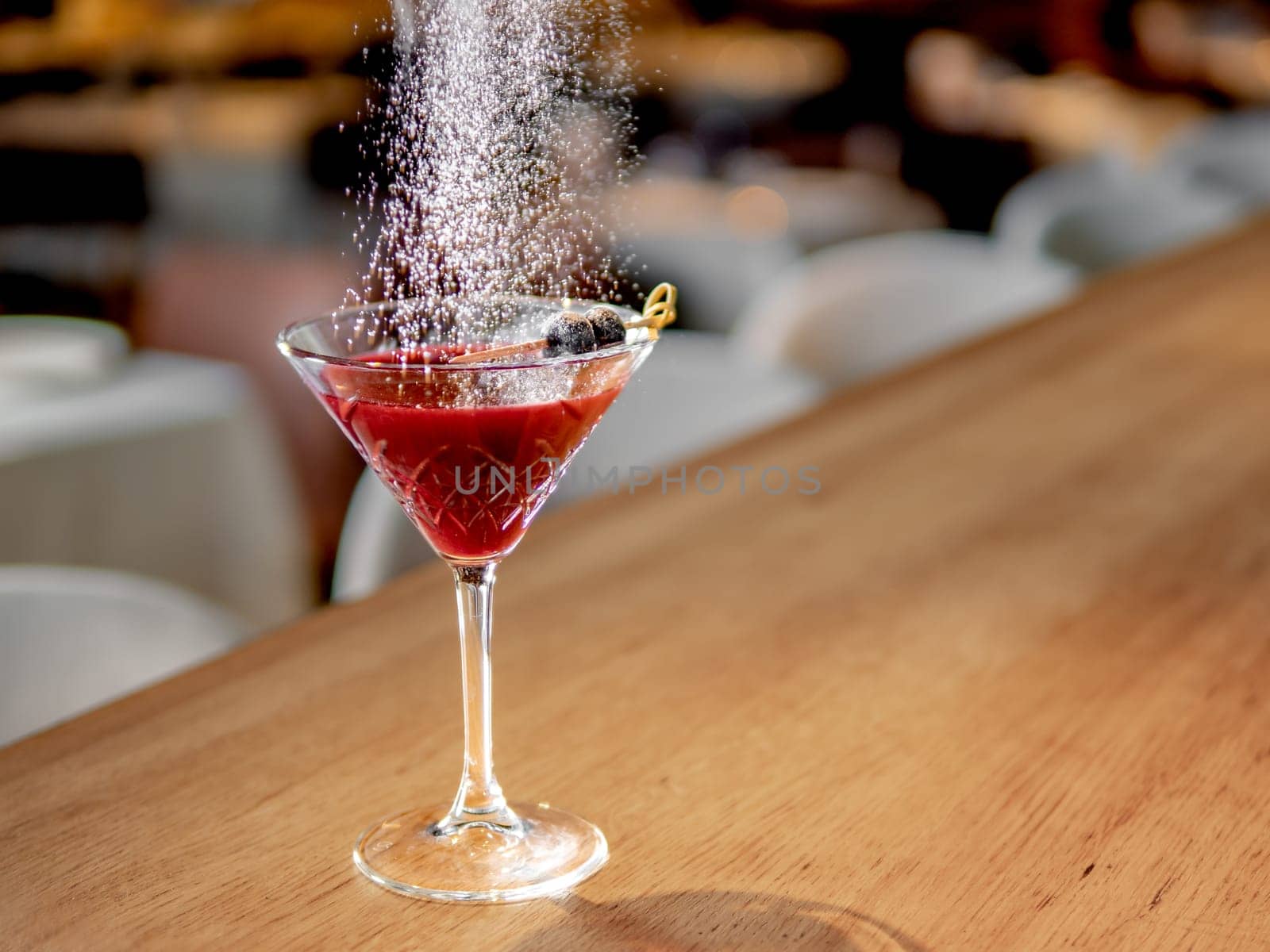 Red cocktail in martini glass on bar counter. White powdered sugar sprinkled on alcohol cocktail decorated black olive. Decorate cold alcoholic cocktail with sugar powder