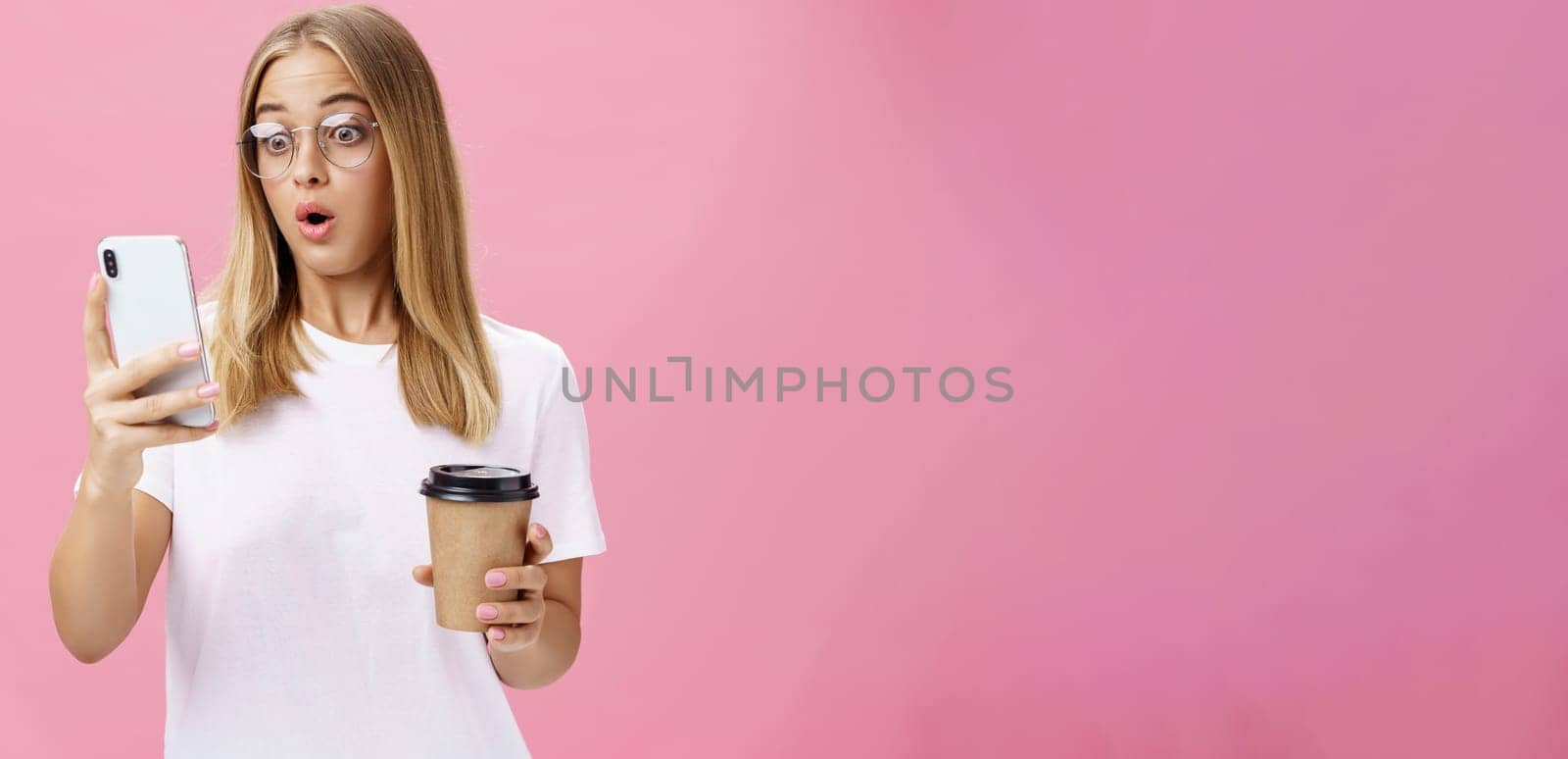 Woman drinking coffee being shocked by received message reacting on stunning news folding lips gasping looking astonished and impressed at smartphone screen holding paper cup, posing over pink wall by Benzoix