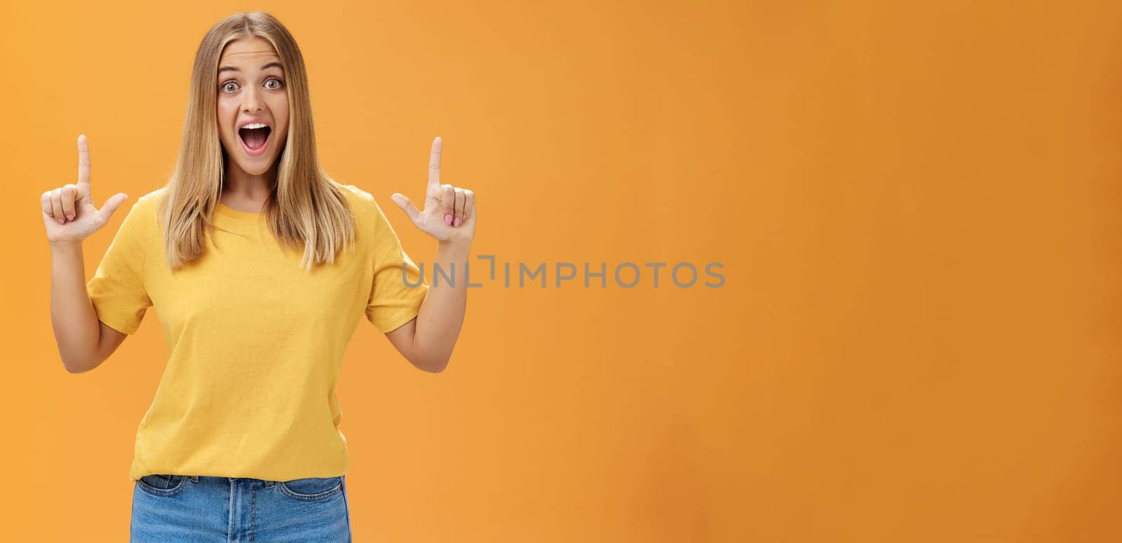 Portrait of joyful surprised and impressed delighted young woman with tanned skin and fair hair opening mouth from amazement and joy raising hands pointing up at awesome copy space against orange wall by Benzoix