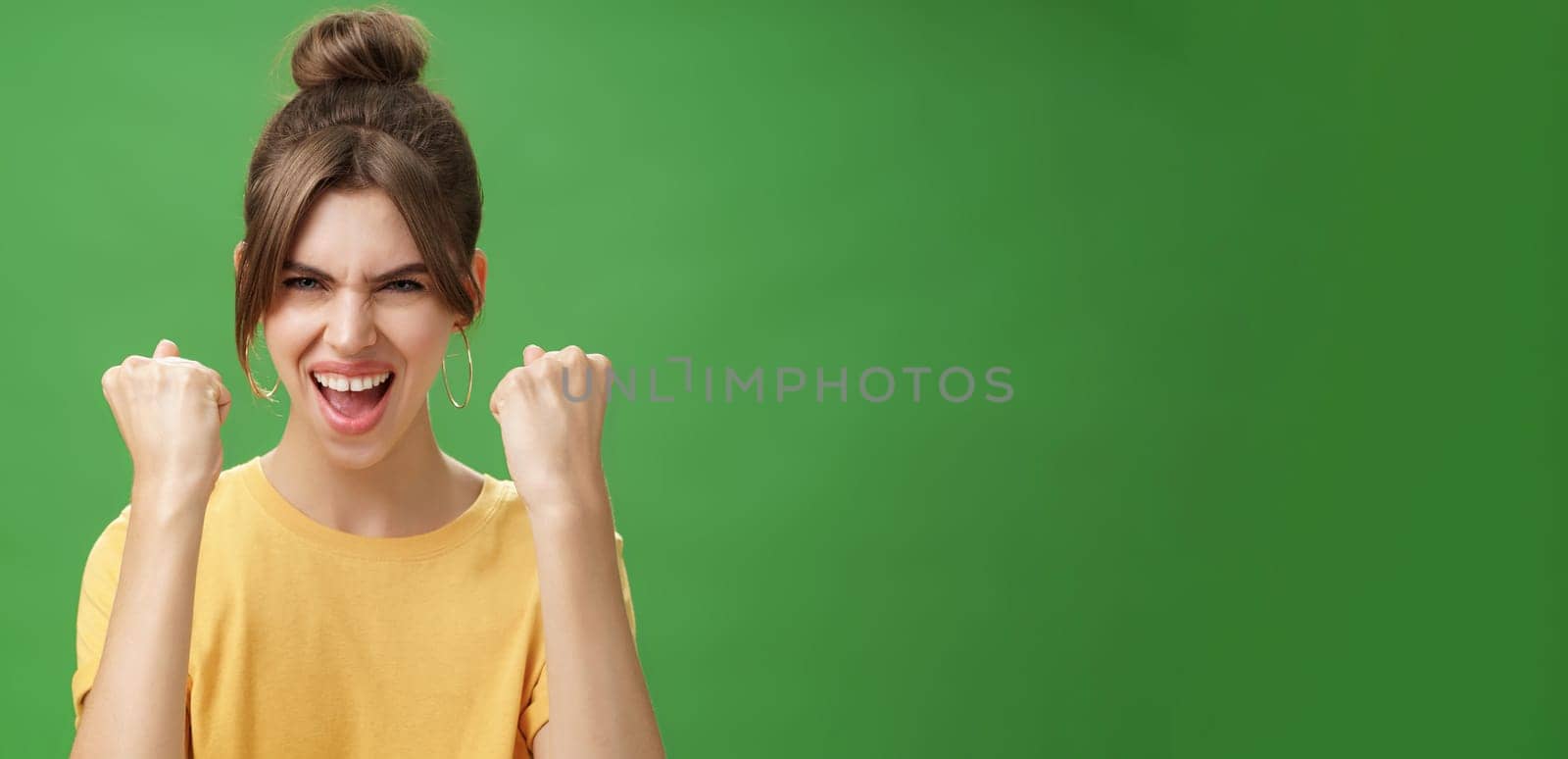 Woman with spirit of winner raising clenched fists smiling excited and supportive cheering being ready for gym excercised boosing confidents with yell looking daring at camera over green wall. Lifestyle.