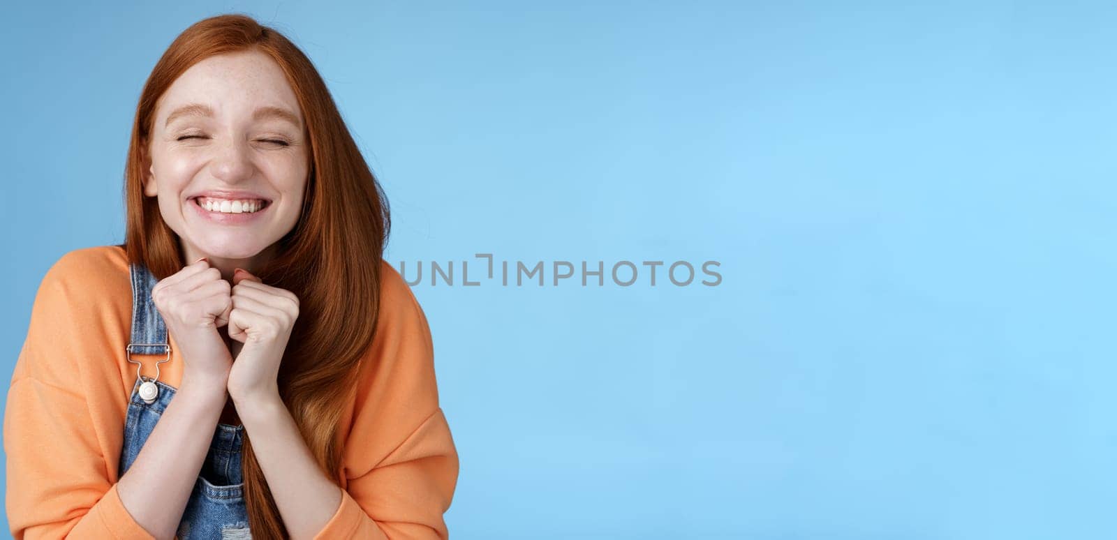 Eager rejoicing thrilled pretty young redhead girl close eyes dreamy smiling receive great result scholarchip triumphing joyfully grinning squeez hands excited, standing blue background very happy by Benzoix