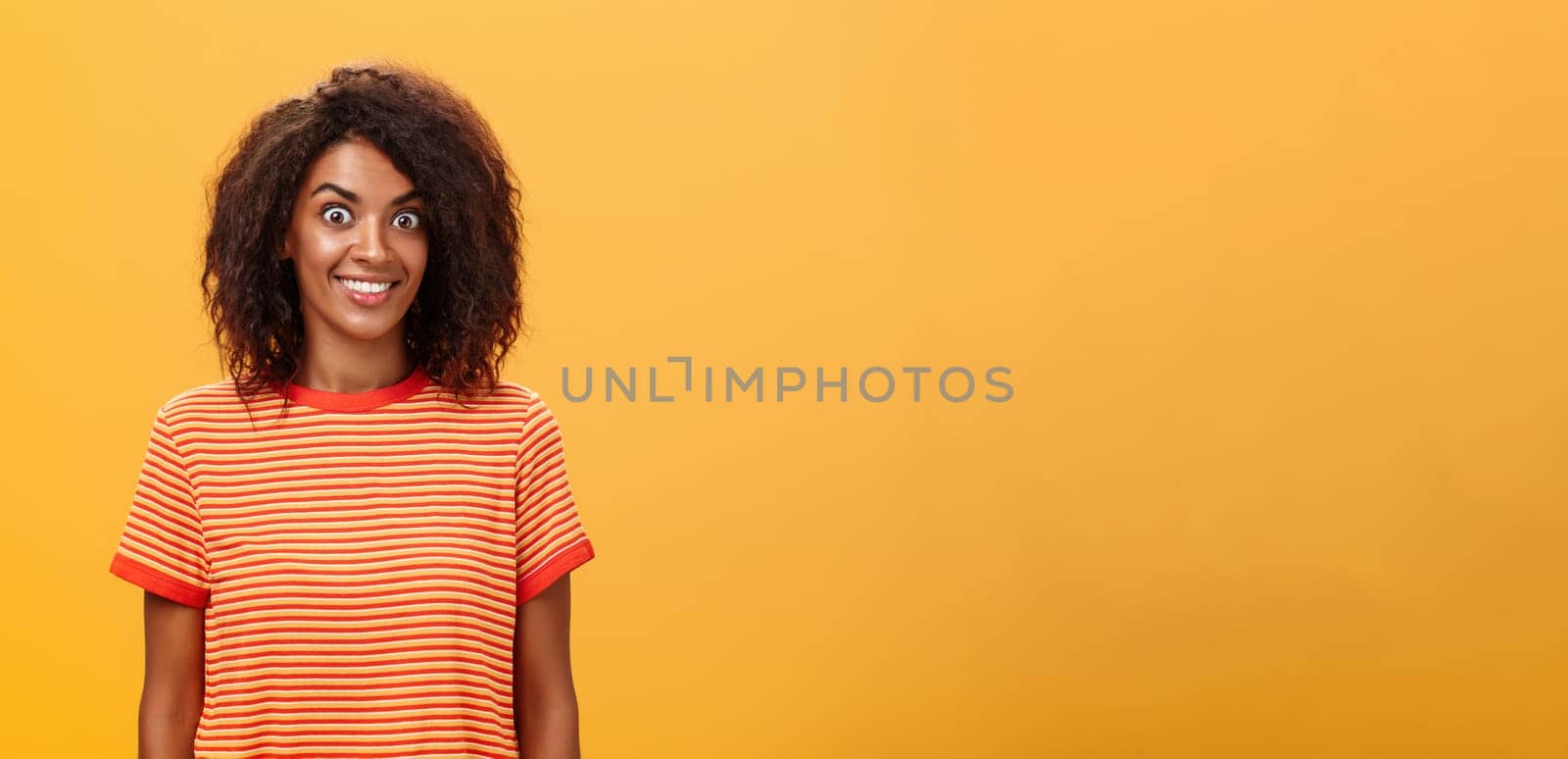 Waist-up shot of amazed and excited charming african american woman with curly hairstyle popping eyes from thrill and joy smiling broadly being surprised by great gift over orange background. Lifestyle.