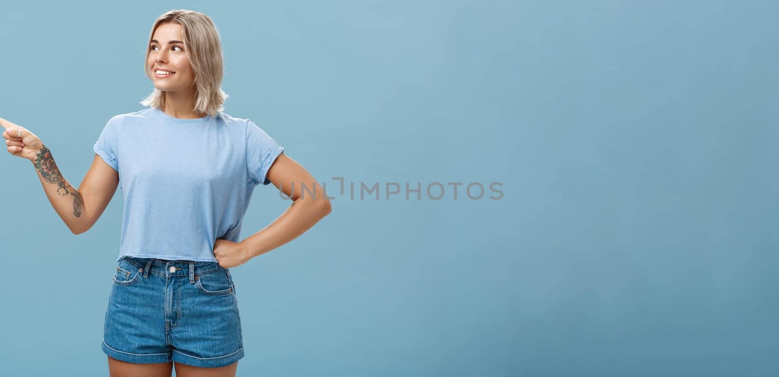 Studio shot of intrigued charming young european female with tanned skin and blond hair holding hand on waist in confident pose looking and pointing left with interest and curiosity. Copy space