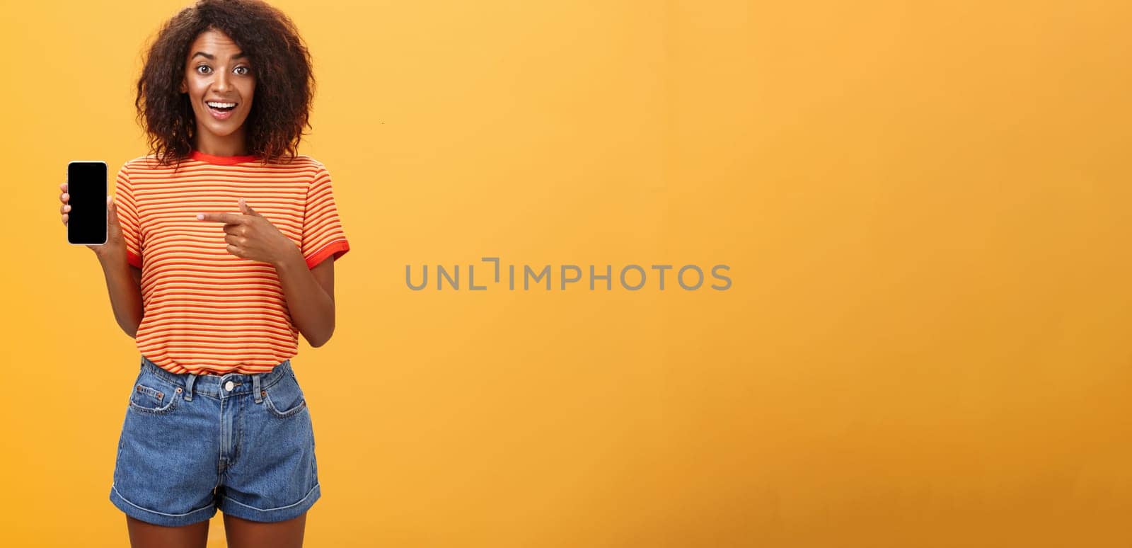 Cute african american stylish woman showing friend new app in smartphone pointing at cellphone screen with index finger smiling thrilled and happy delighted with cool device purchase over orange wall by Benzoix