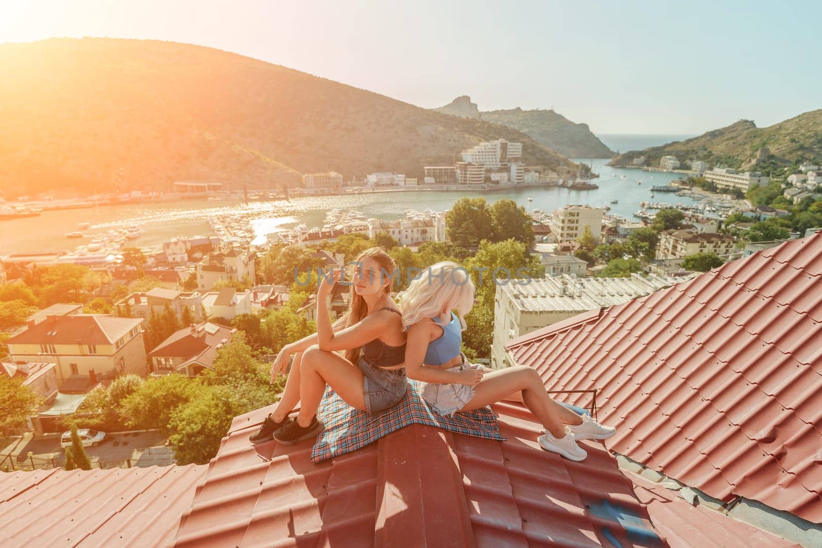 Two women sitting on a red roof, enjoying the view of the town and the sea. Rooftop vantage point. In the background, there are several boats visible on the water, adding to the picturesque scene. by Matiunina
