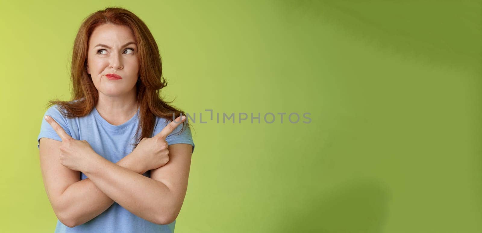 Time make right choice. Perplexed hesitant redhead middle-aged female customer deciding between few variants cross arms body pointing sideways look left smirking unsure doubting choosing product.