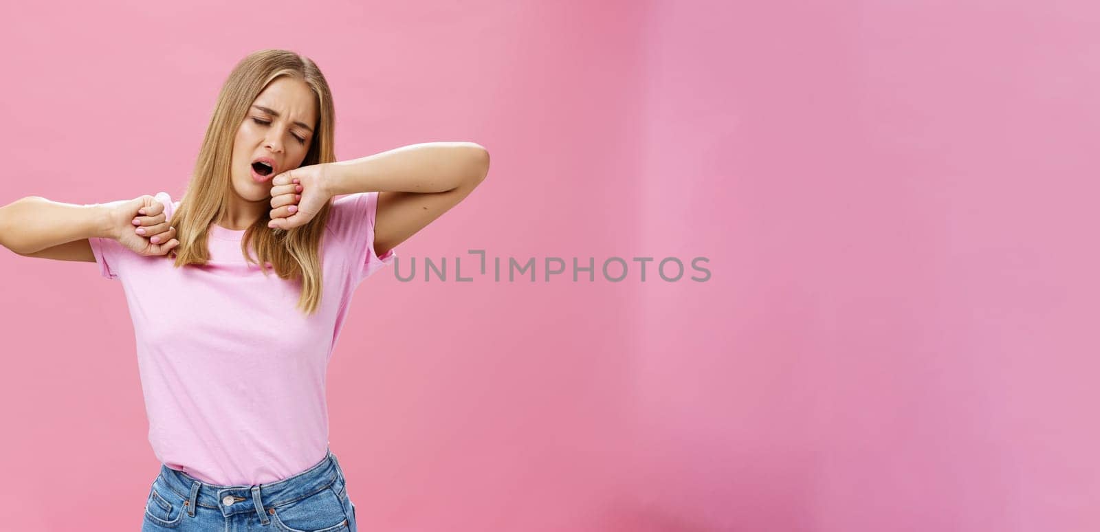 Too tired to work today. Lazy and exhausted attractive young female student doing homework all night yawning with closed eyes while stretching hands from after tiresome project over pink wall by Benzoix