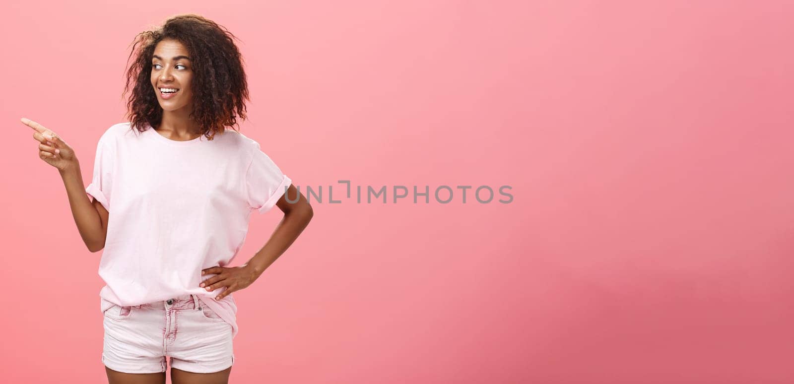 Girl checking out nice body of guy passing by. Charming confident dark-skinned female with curly hairstyle holding hand on hip looking and pointing left discussing cool copy space over pink wall by Benzoix
