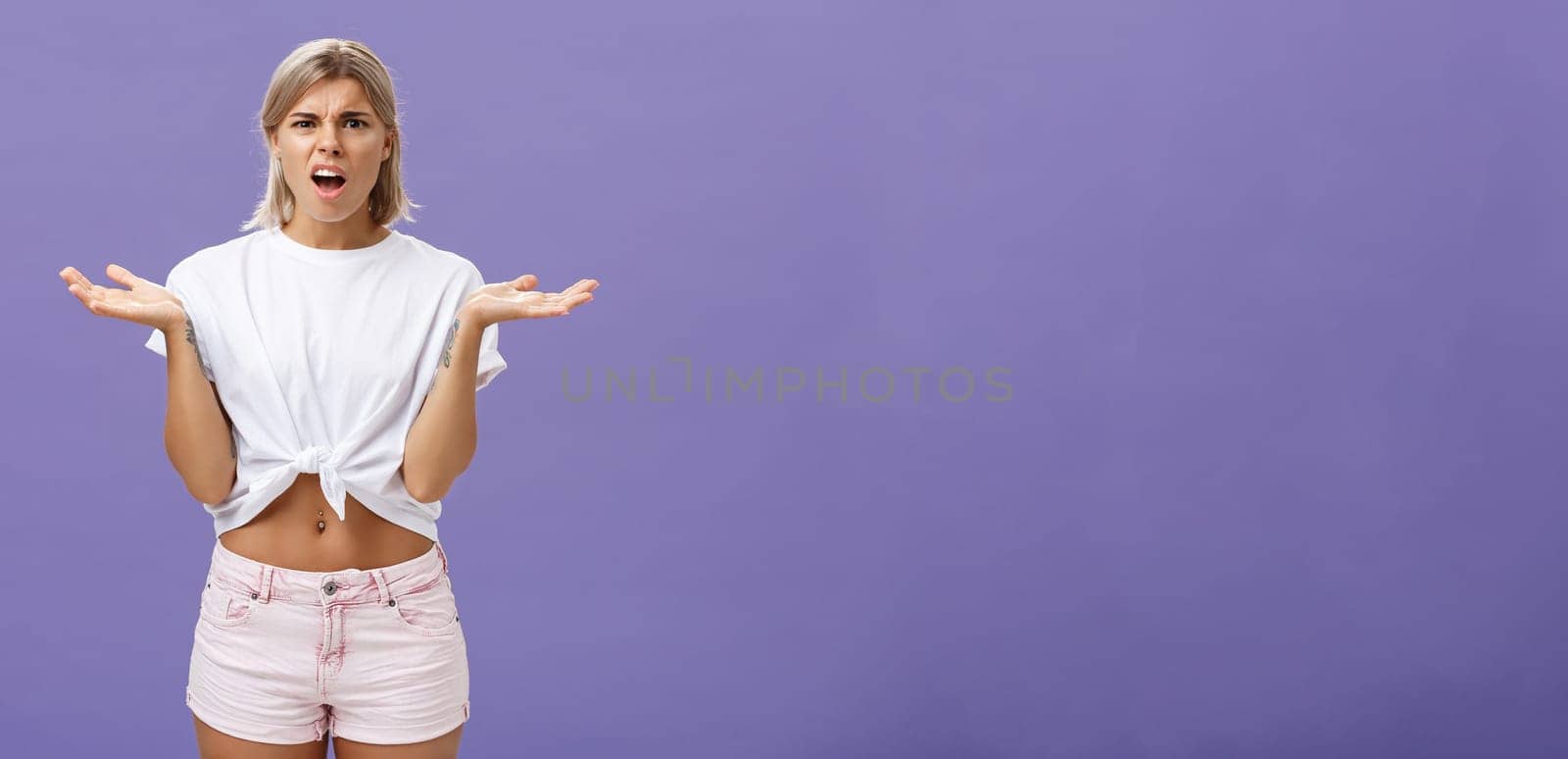 Perplexed and disappointment good-looking blonde female student in white t-shirt and pink shorts frowning shrugging with spread hands near shoulders saying what the hell over purple wall by Benzoix