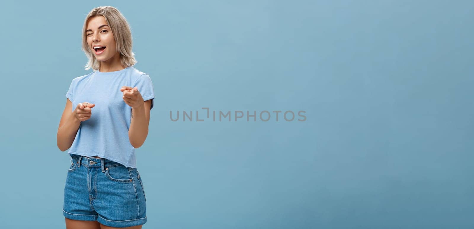 Hey I got offer for you. Portrait of attractive self-assured and playful stylish blond female with tanned skin in denim shorts and summer t-shirt winking and pointing at camera over blue background by Benzoix