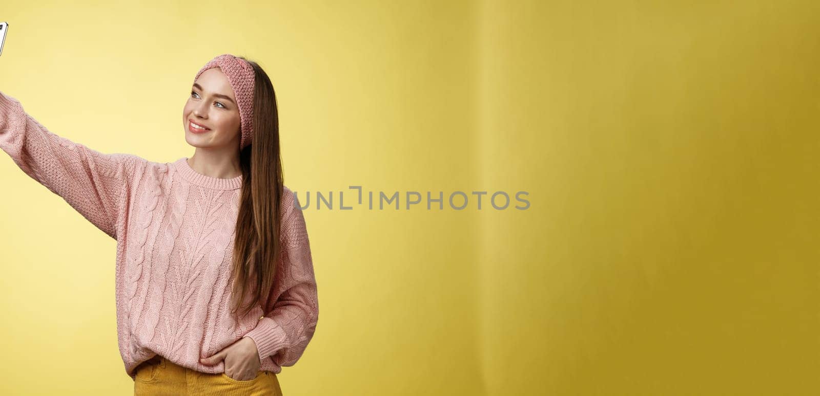 Popular glamour young female internet lifestyle blogger taking selfie on new smartphone extending arm taking picture herself against yellow background smiling at cellphone screen, posing cheeky by Benzoix