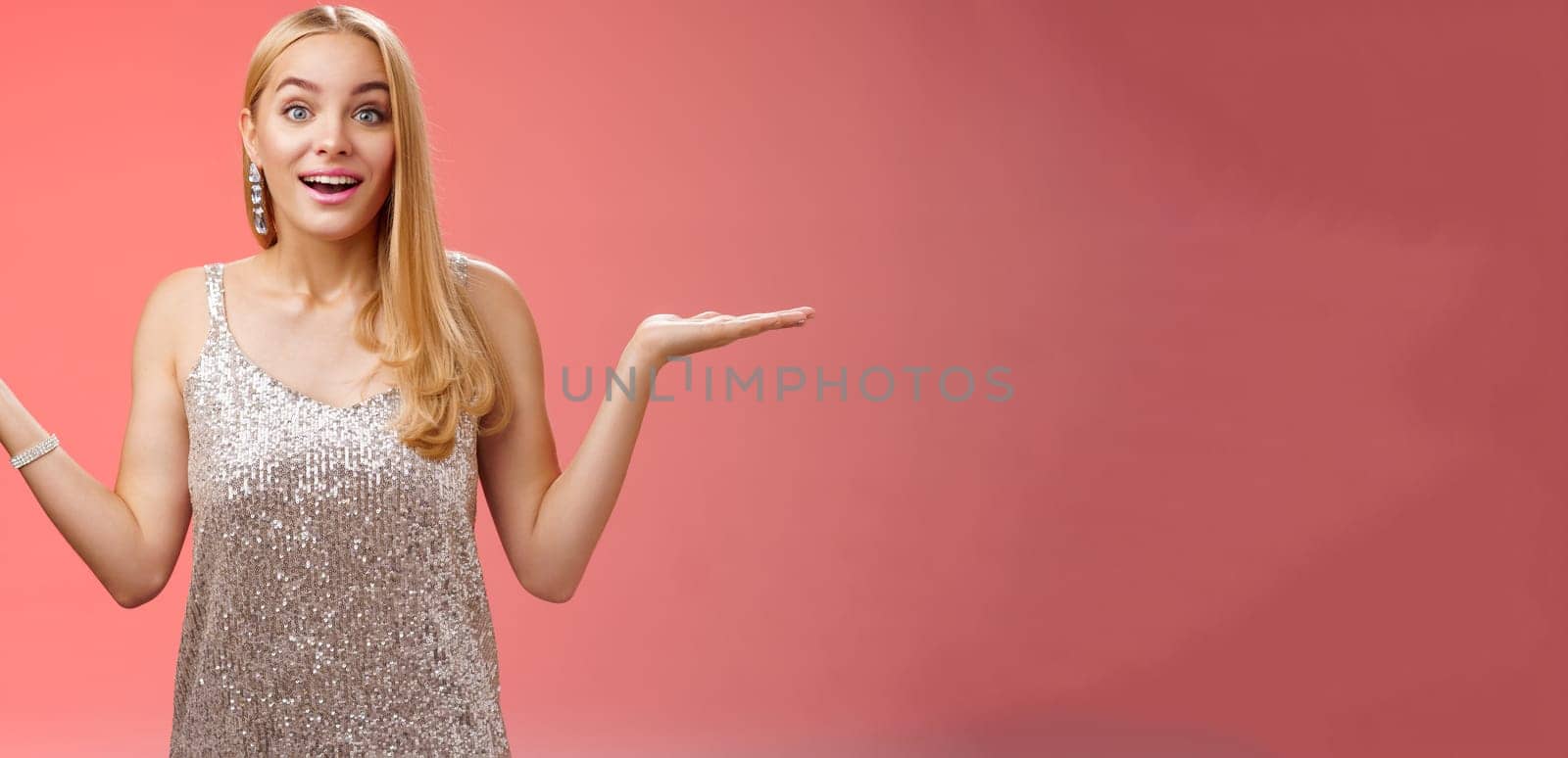 Carefree fascinated young blond girl 20s in silver glittering evening party dress raising hands sideways weighing shrugging smiling amused search right choice, deciding what do red background by Benzoix