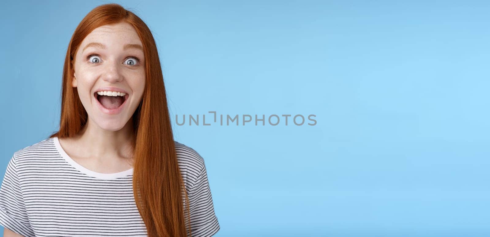 Surprised pleased happy impressed redhead european girl 20s reacting amused wide eyes look admiration joy receive incredible offer standing excited express thrill upbeat feelings, blue background.
