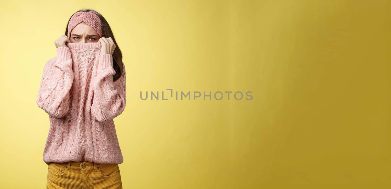 Sad silly, gloomy cute young girl hiding face in sweater collar pulling clothes on nose frowning displeased, unhappy, looking indignant and reluctant standing disappointed over yellow background.