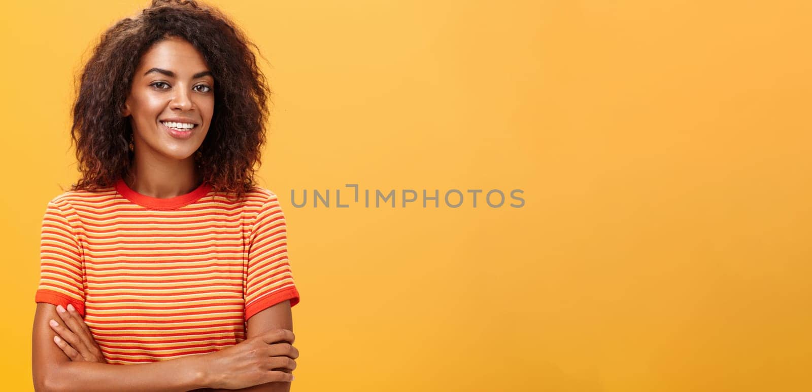 Waist-up shot of creative ambitious good-looking female coworker in trendy striped t-shirt holding hands crossed on chest smiling broadly at camera friendly and self-assured over orange background by Benzoix