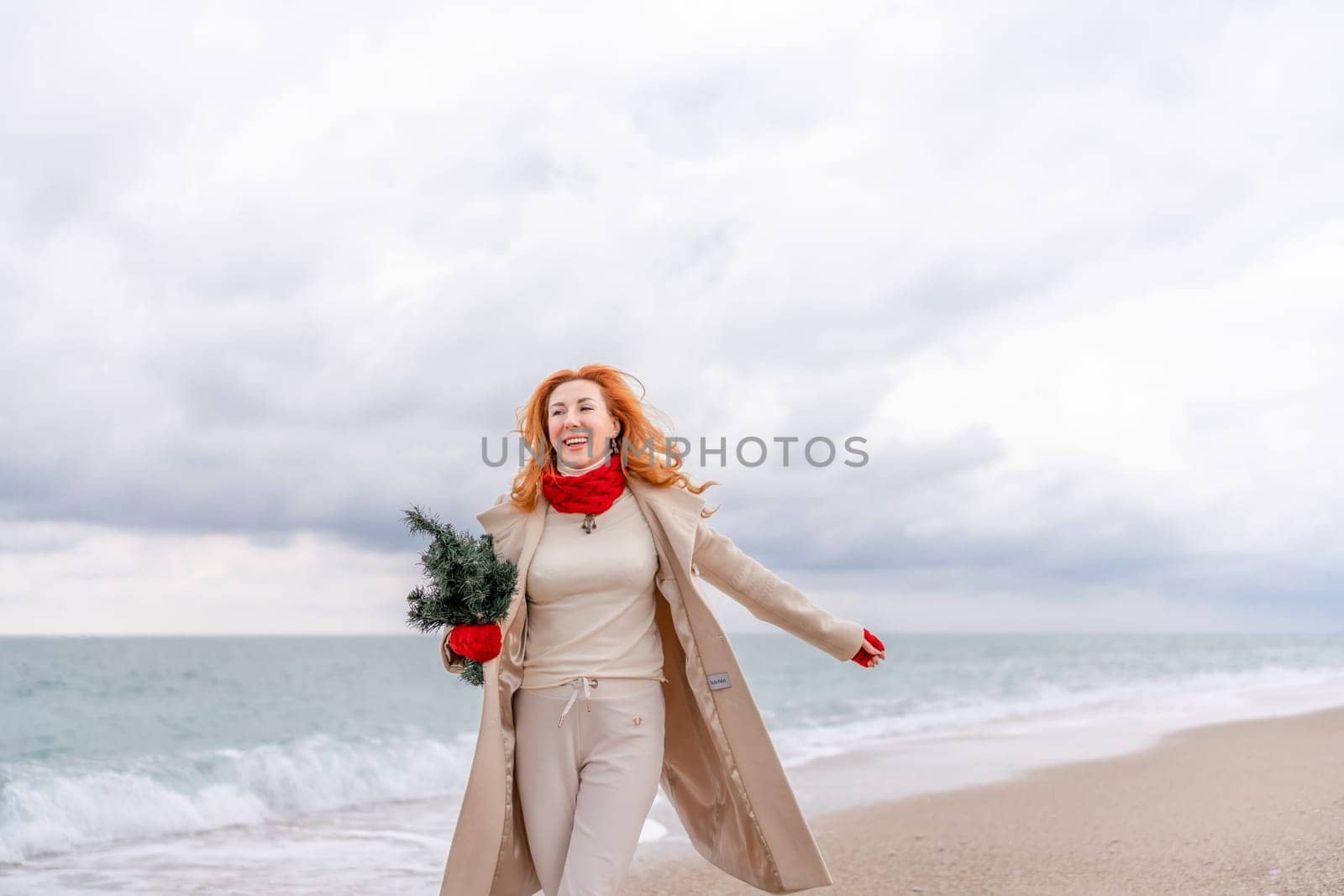 Redhead woman Christmas tree sea. Christmas portrait of a happy redhead woman walking along the beach and holding a Christmas tree in her hands. Dressed in a light coat, white suit and red mittens