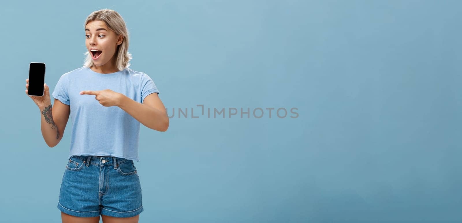 Look at this awesome smartphone. Impressed enthusiastic good-looking female blonde with cool tattoo on arm dropping jaw from thrill and excitement pointing at device screen over blue background by Benzoix