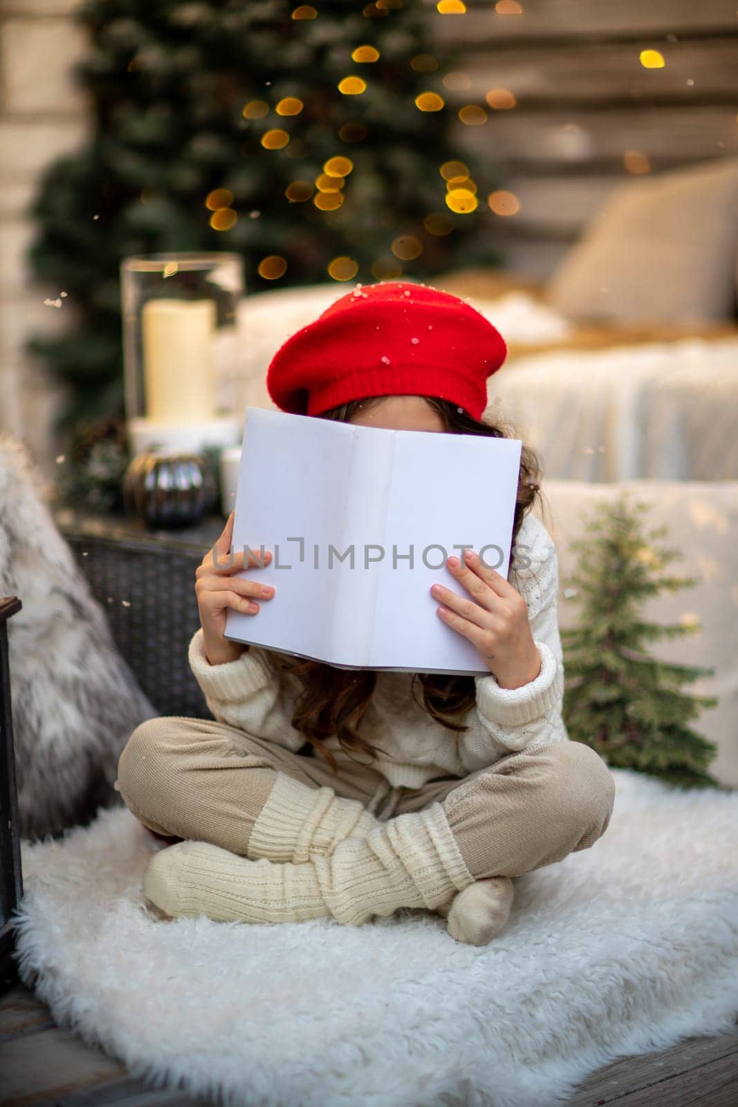 Girl with a book under the Christmas tree. Portrait of a cute girl in a red beret, she sits on the floor and covers her face with a book. Children and New Year mood