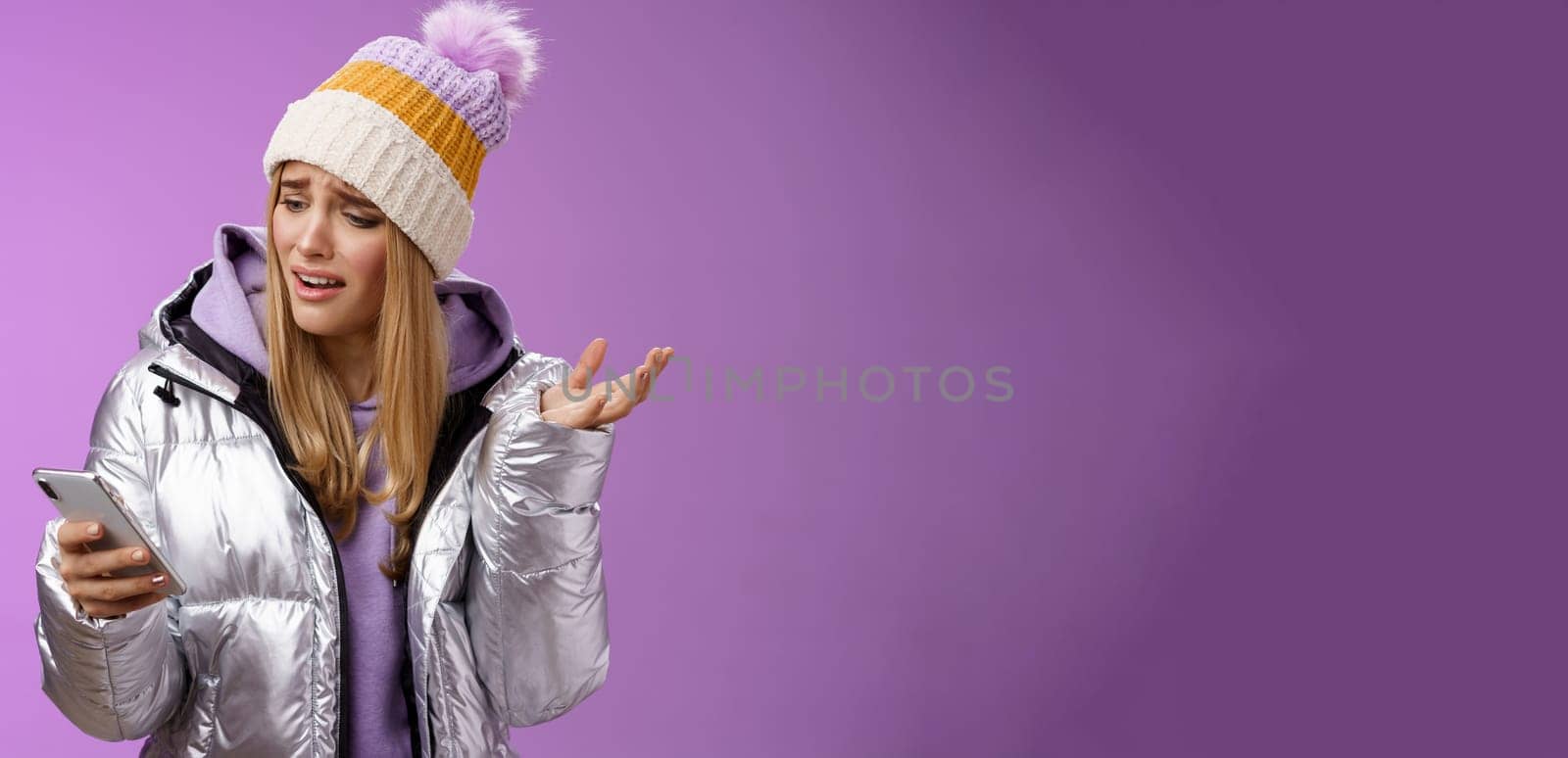 Upset disappointed attractive whining gloomy blond girl in silver jacket standing outside hat holding smartphone shrugging raising hand dismay complaining slow mobile internet, purple background.