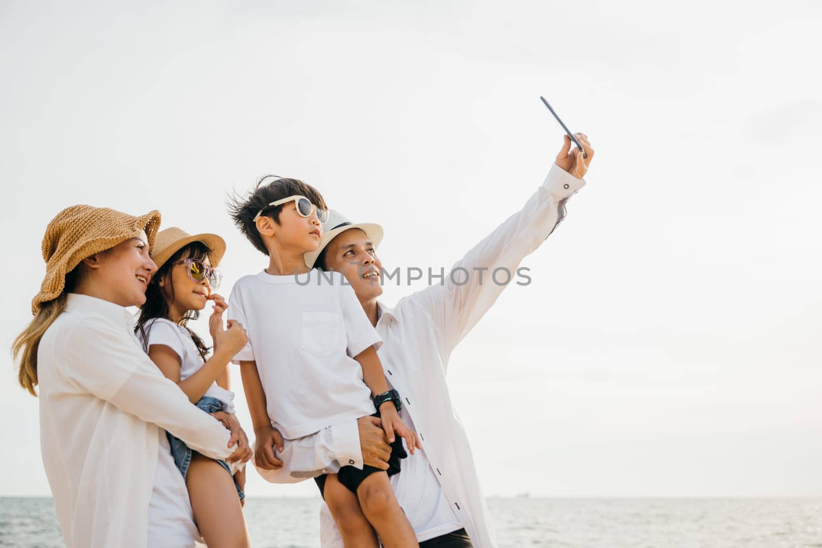 Beachside family fun as they take a cheerful selfie together near the sea. Laughter and joy unfold creating a playful and memorable moment of togetherness during a summer travel adventure. by Sorapop