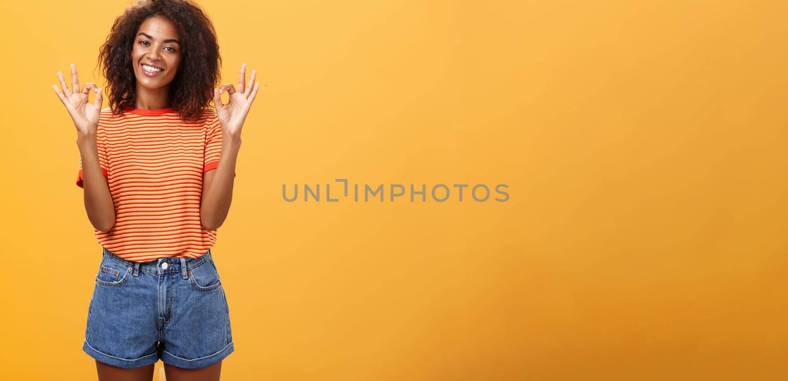 Chill attractive satisfied african american female customer picking new outfit being delighted and pleased with cool staff raising hands in okay or awesome gesture smiling happily over orange wall by Benzoix
