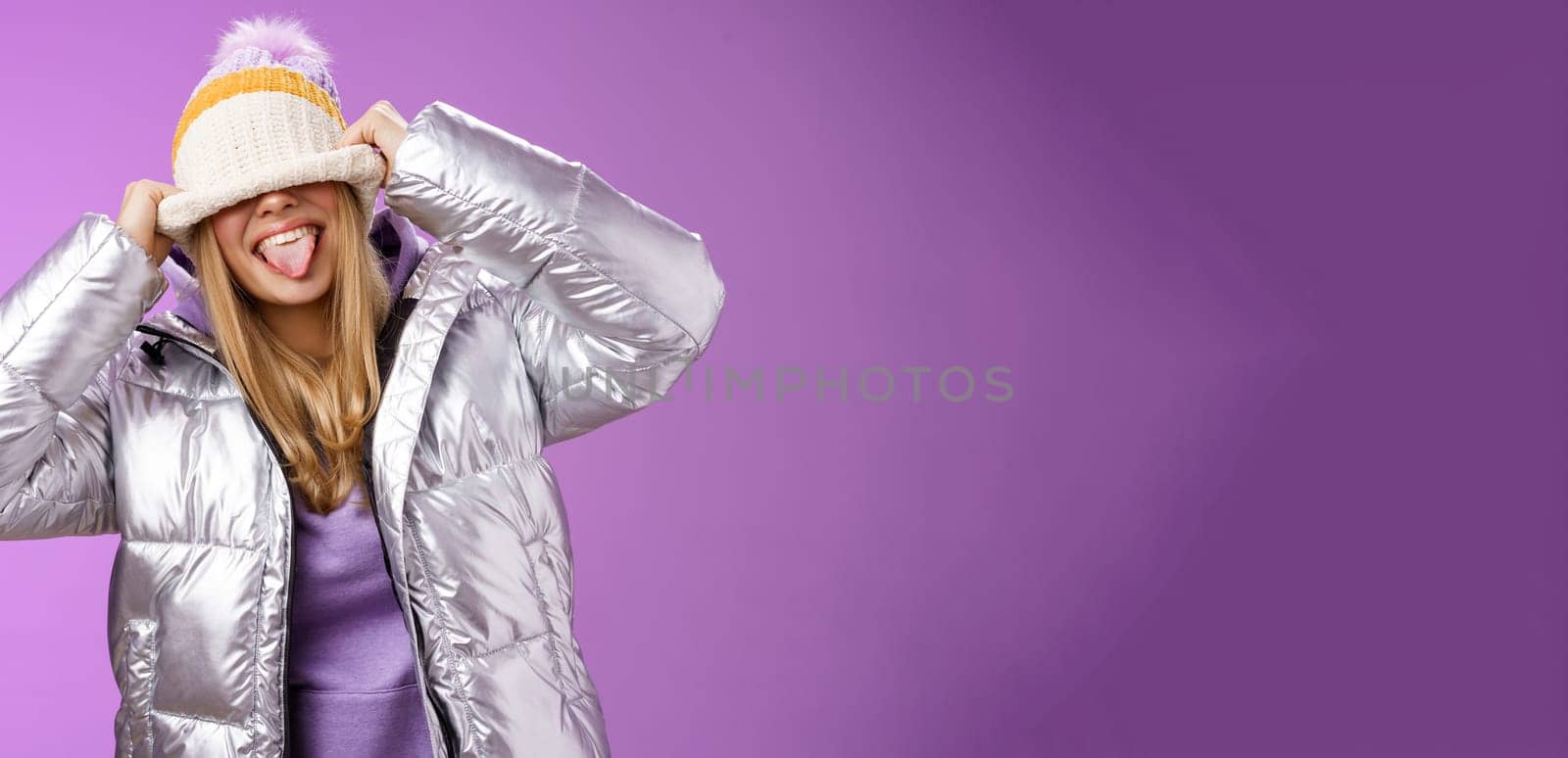 Playful carefree funny blond girl having fun fool around careless what people thing acting immature joyfully pulling hat eyes sticking tongue smiling standing silver jacket purple background by Benzoix