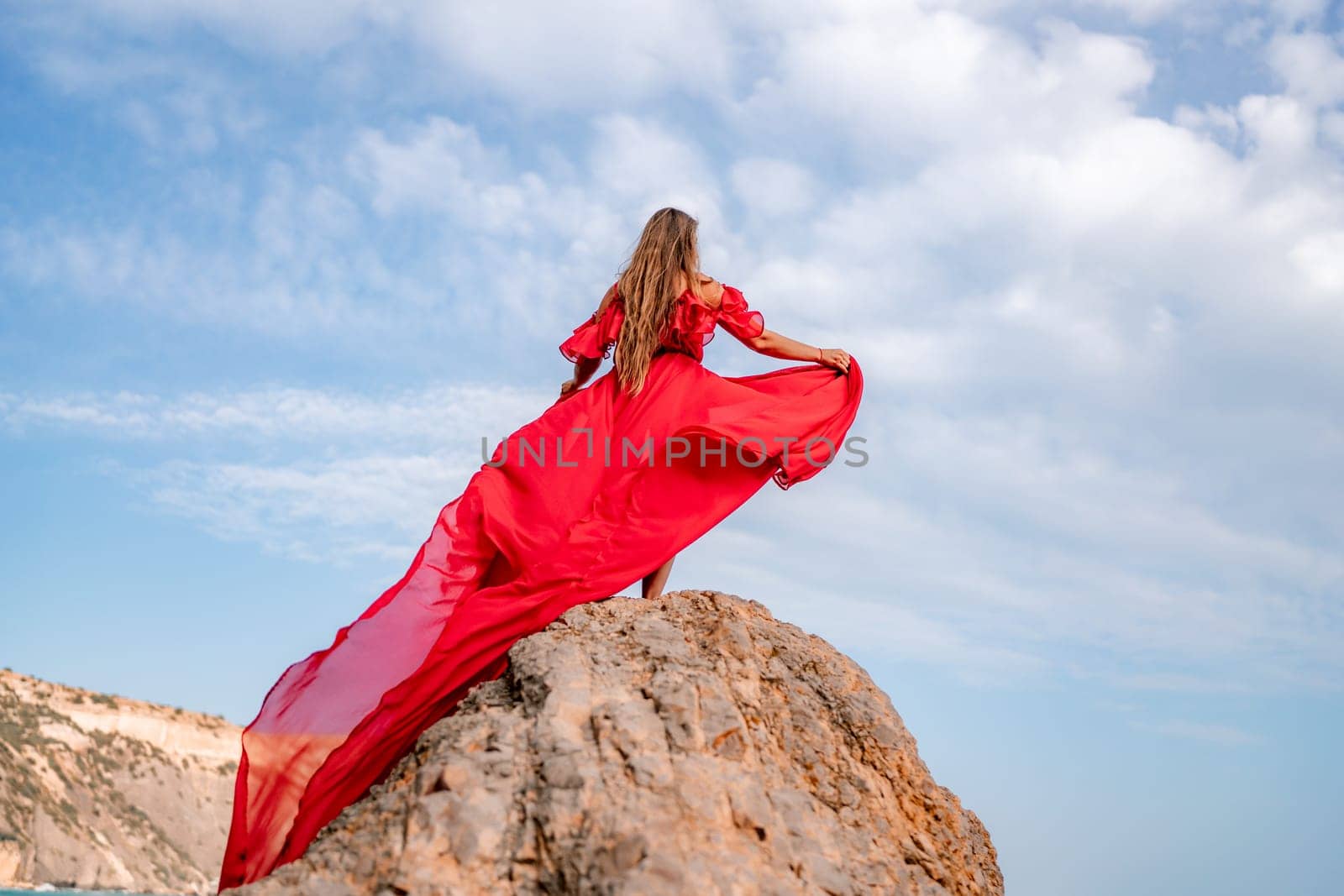woman sky red dress. Woman with long hair on a sunny seashore in a red flowing dress, back view, silk fabric waving in the wind. Against the backdrop of the blue sky and mountains on the seashore