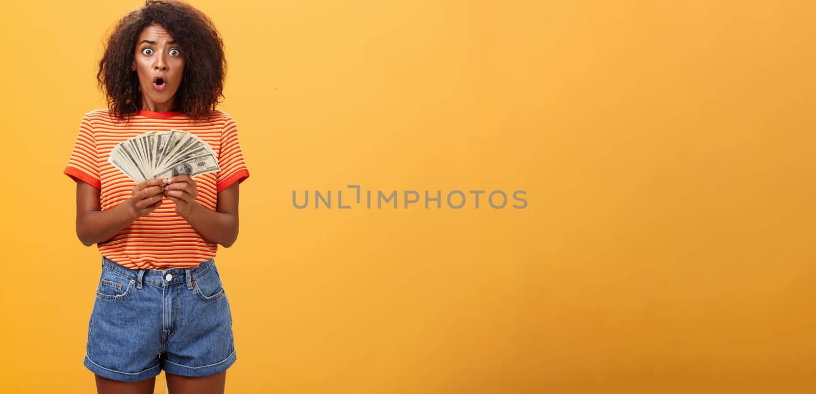 Woman shocked finding lots of cash in safe. Portrait of surprised speechless good-looking dark-skinned female with curly haircut folding lips gasping holding money posing over orange background by Benzoix