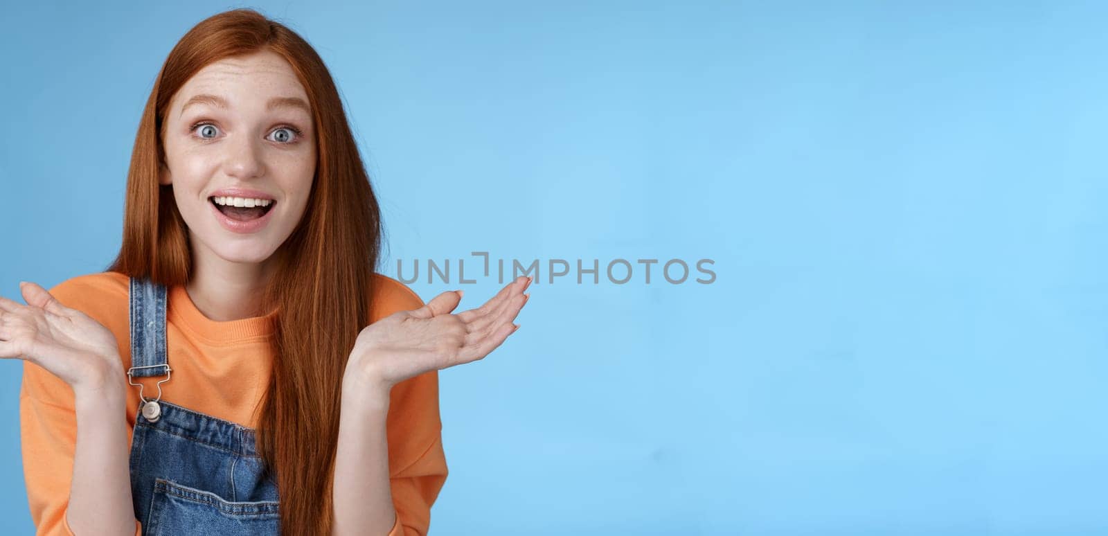 Amused surprised glad young redhead girl speechless happy see friend came back hometown wide eyes impressed grinning raise hands sideways full disbelief standing blue background joyful by Benzoix