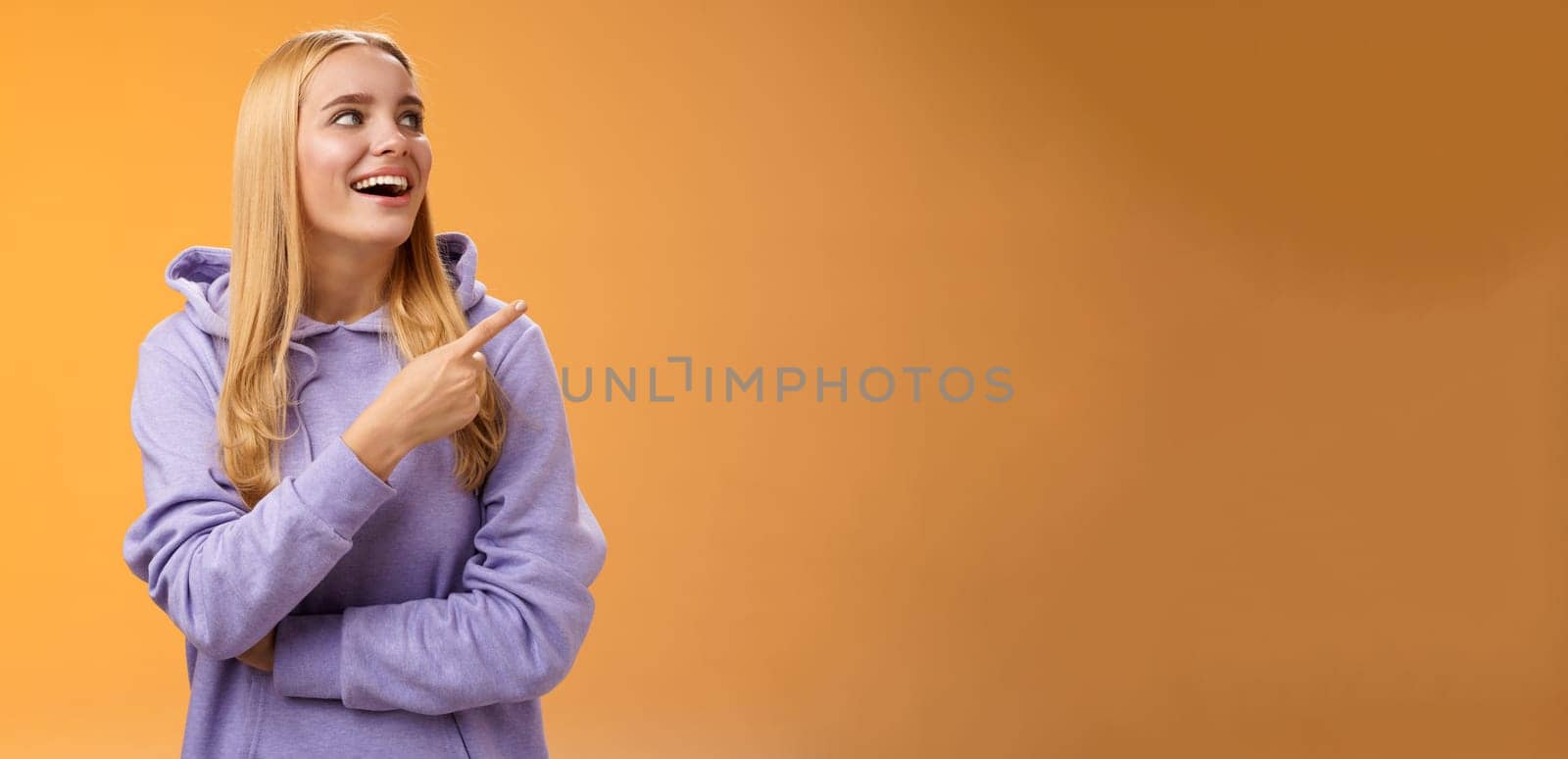 Amused carefree joyful blond european woman in hoodie pointing looking right pleased smiling happily enjoying interesting fascinating peromance making choice in store shopping, orange background by Benzoix
