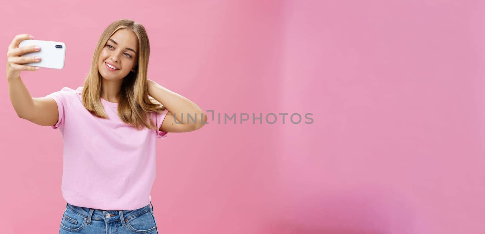 Female beauty blogger taking selfie post new look online. Portrait of charming tanned young woman in t-shirt touching hair gently pulling hand with smartphone near face taking photo over pink wall by Benzoix