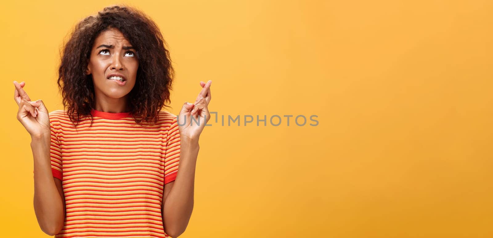 Waist-up shot of hopeful concerned and anxious charming dark-skinned woman with afro hairstyle in trendy striped t-shirt biting lip nervously looking up crossing fingers for good luck while praying. Lifestyle.