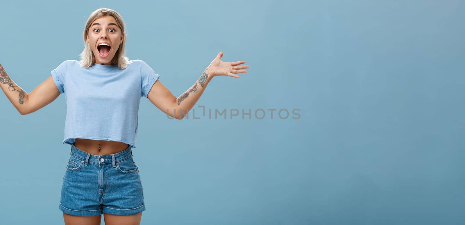 Girl overwhelmed with positive emotions and happiness being surprised rejoicing and screaming from joy spreading palms aside opening mouth from amazement standing delighted over blue wall by Benzoix