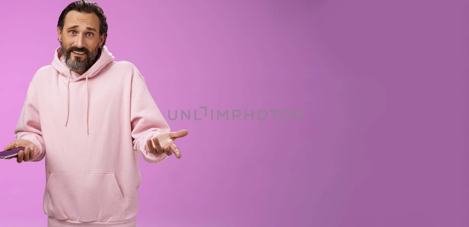 So what. Confused questioned adult bearded stylish guy in pink hoodie shrugging hands spread sideways dismay holding smartphone look clueless unaware camera not know how deal annoying phone calls by Benzoix