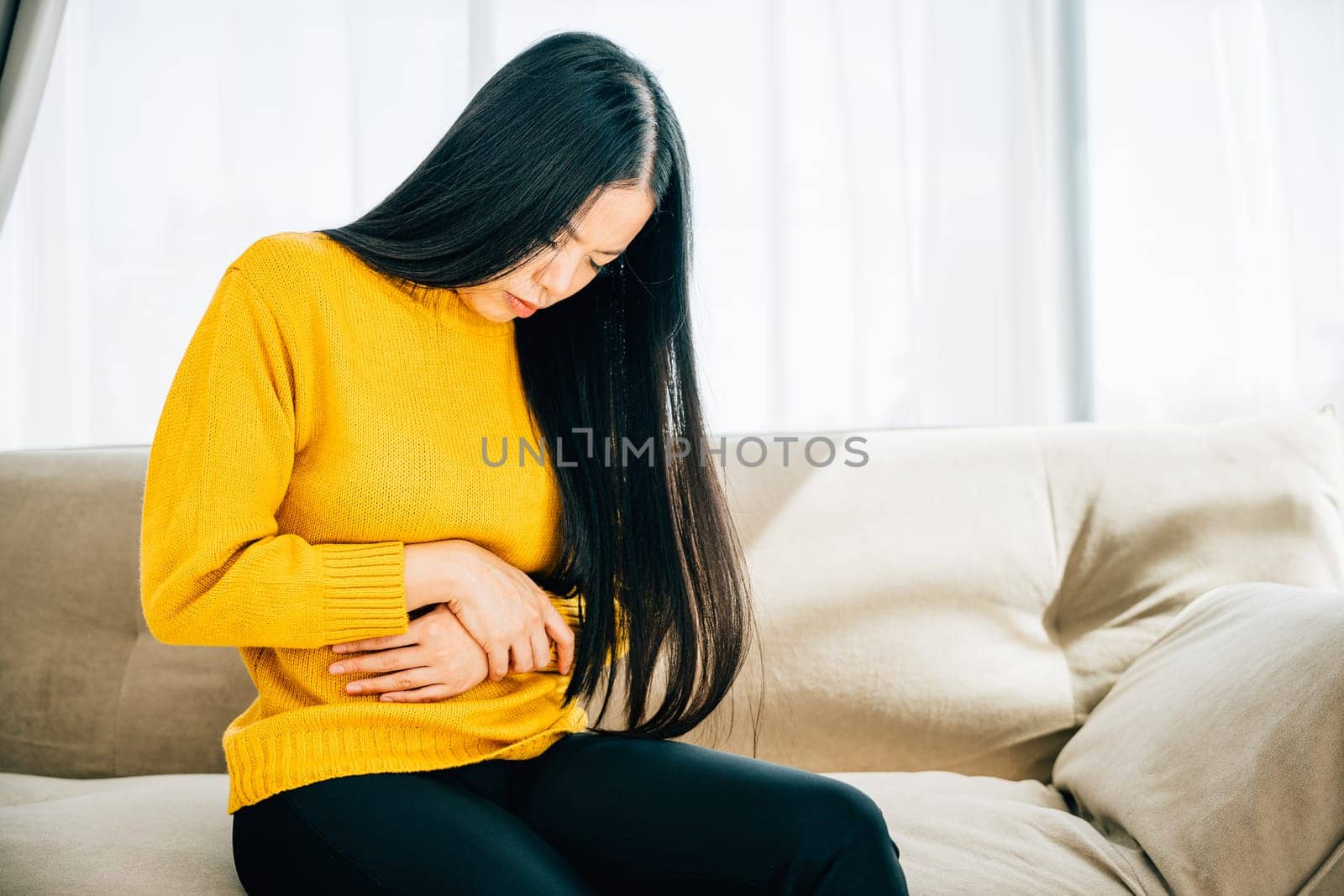 Depicting stomachache, Asian woman on home couch suffers from abdominal pain. Emphasizing discomfort symptoms and the need for medical care for stomachache. Health care abdomen bloating concept