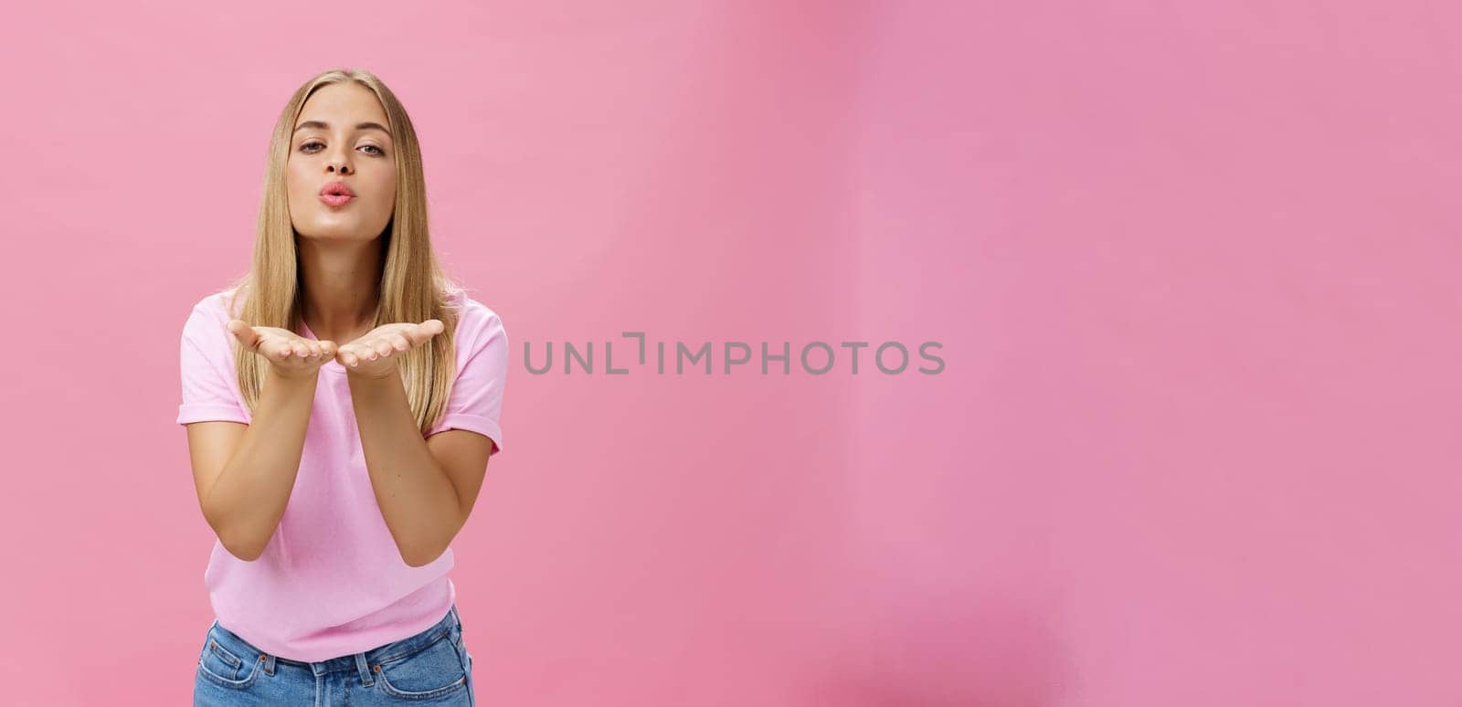 Feminine and flirty charming pretty girl with tanned skin and fair straight hair holding palms near folded lips sending air kiss at camera with sensual expression posing over pink background. Lifestyle.