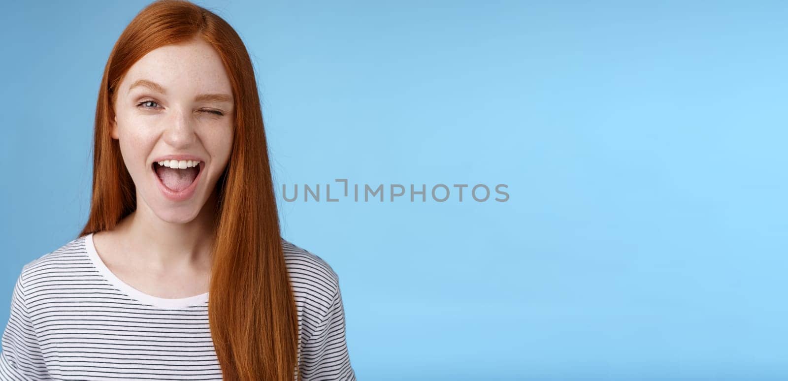 Cheeky good-looking flirty young gorgeous redhead girlfriend winking sassy smiling delighted open mouth hinting have interesting suggestion keep secret standing amused blue background by Benzoix