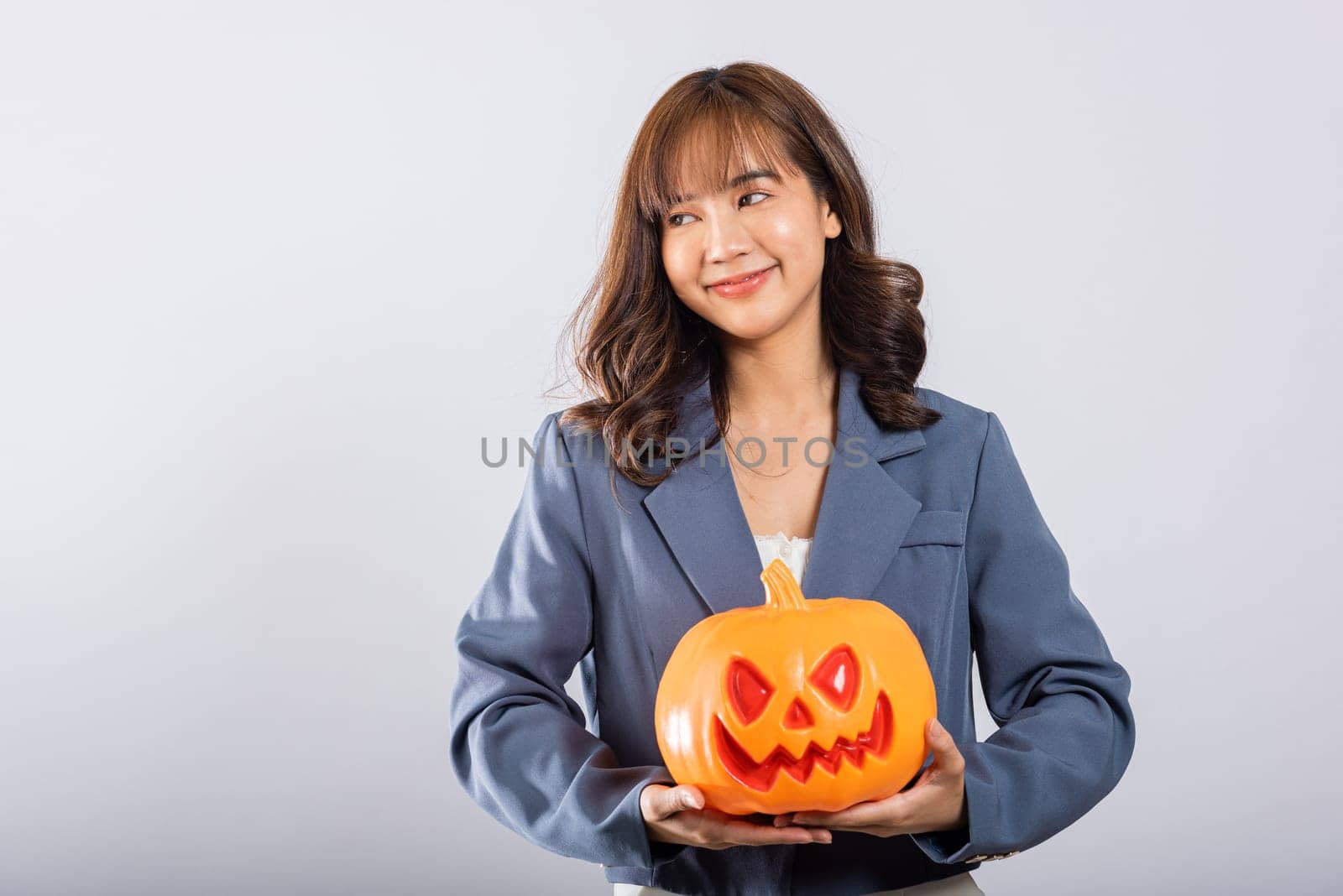 A charming young woman's portrait captures her delight as she holds an orange pumpkin, her joy mirrored by a collection of ghost pumpkins in this studio shot, all set against a white background.