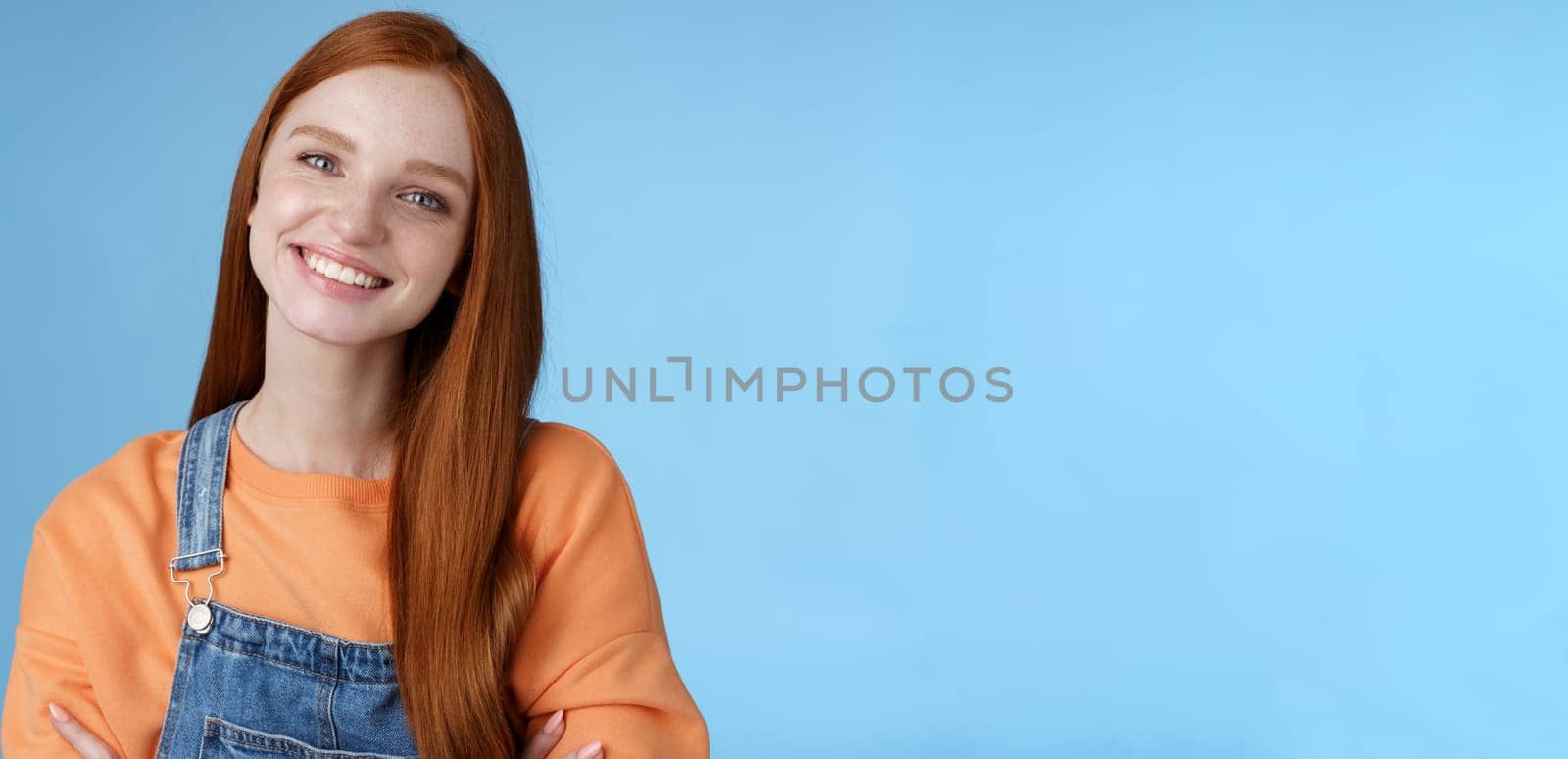 Charismatic kind pleasant redhead girl blue eyes smiling friendly listen politely customer standing blue background tilting head amused grinning cross hands chest professional confidence pose.