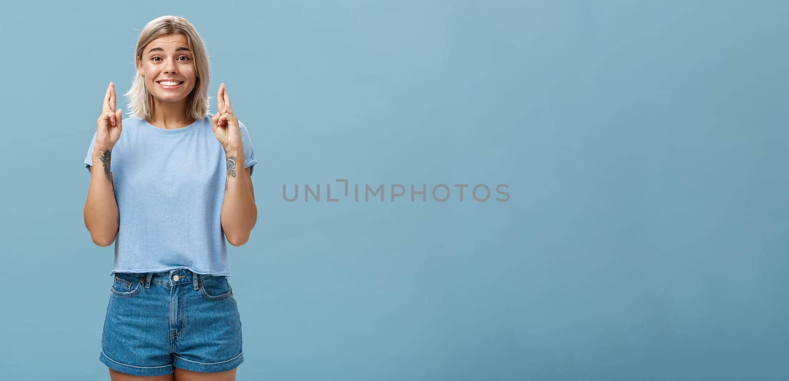 Hope dream come true soon. Portrait of emotive happy and optimistic attractive young european woman with blond hair and tanned skin crossing fingers for good luck and smiling over blue wall by Benzoix