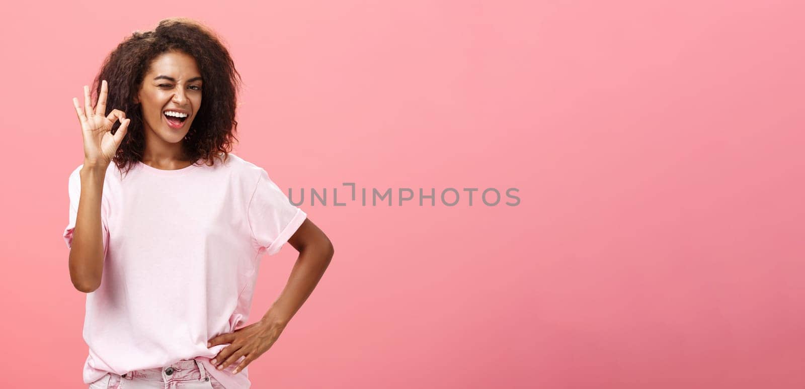 Girl got us covered. Charming charismatic and confident african american skillful woman in stylish outfit holding hand on waist showing okay gesture and winking assured at camera over pink wall.