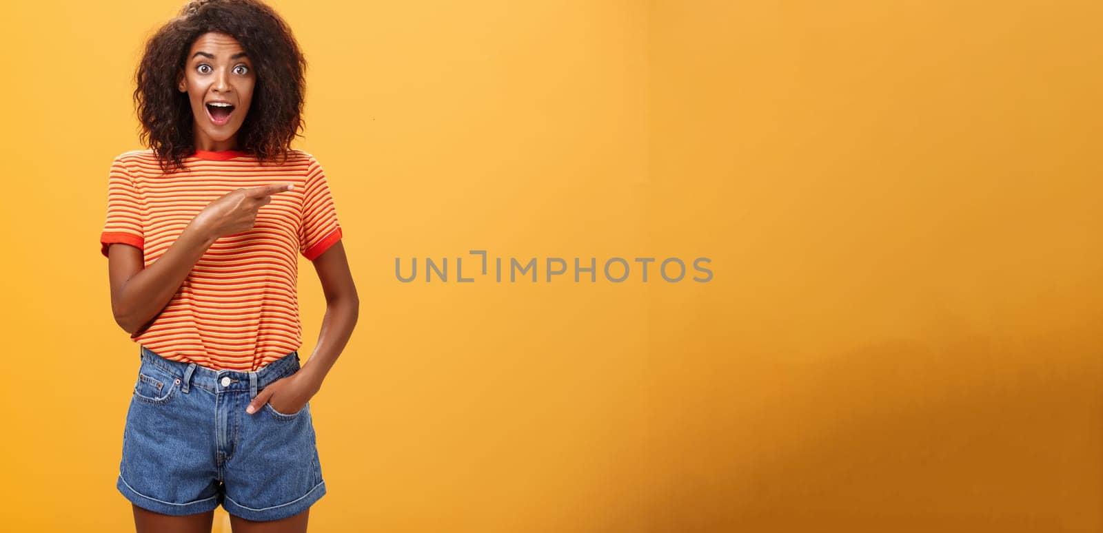 Hey awesme copy space there. Portrait of impressed and surprised enthusiastic young african american female with afro hairstyle pointing right astonished looking energized and curious over orange wall by Benzoix